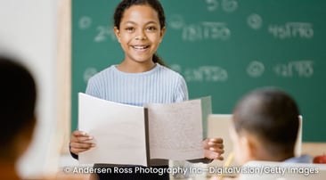 Student standing in front of a class with presentation materials in hand.