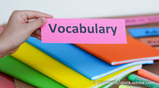 A hand holding a piece of paper with vocabulary written on it and books and supplies in the background.